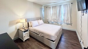 A bedroom with a bed, bedside table, lamp, curtains, and a television mounted on the wall.