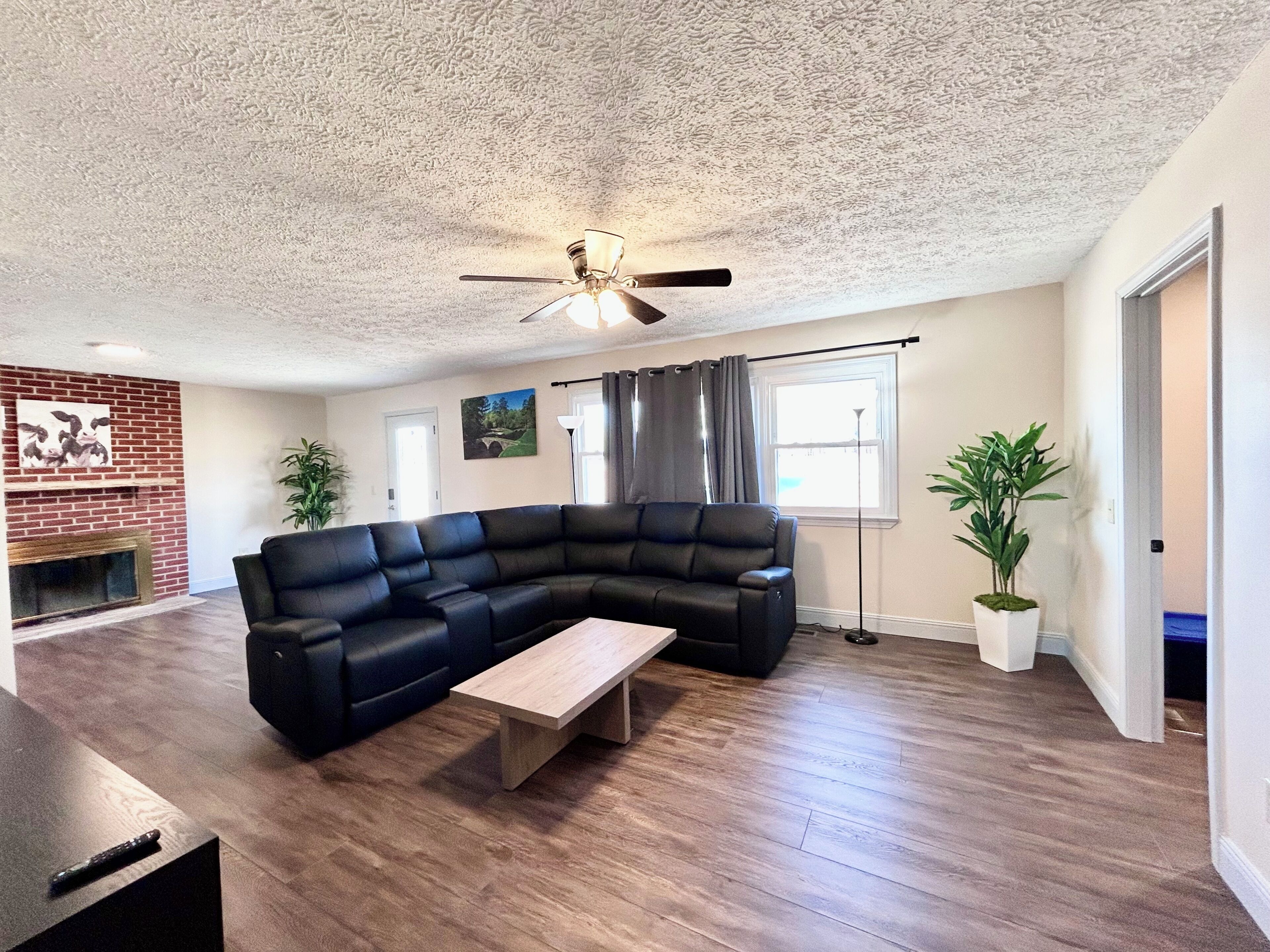 A living room with a sectional sofa, a wooden coffee table, a ceiling fan, a brick fireplace, and a large window with curtains.