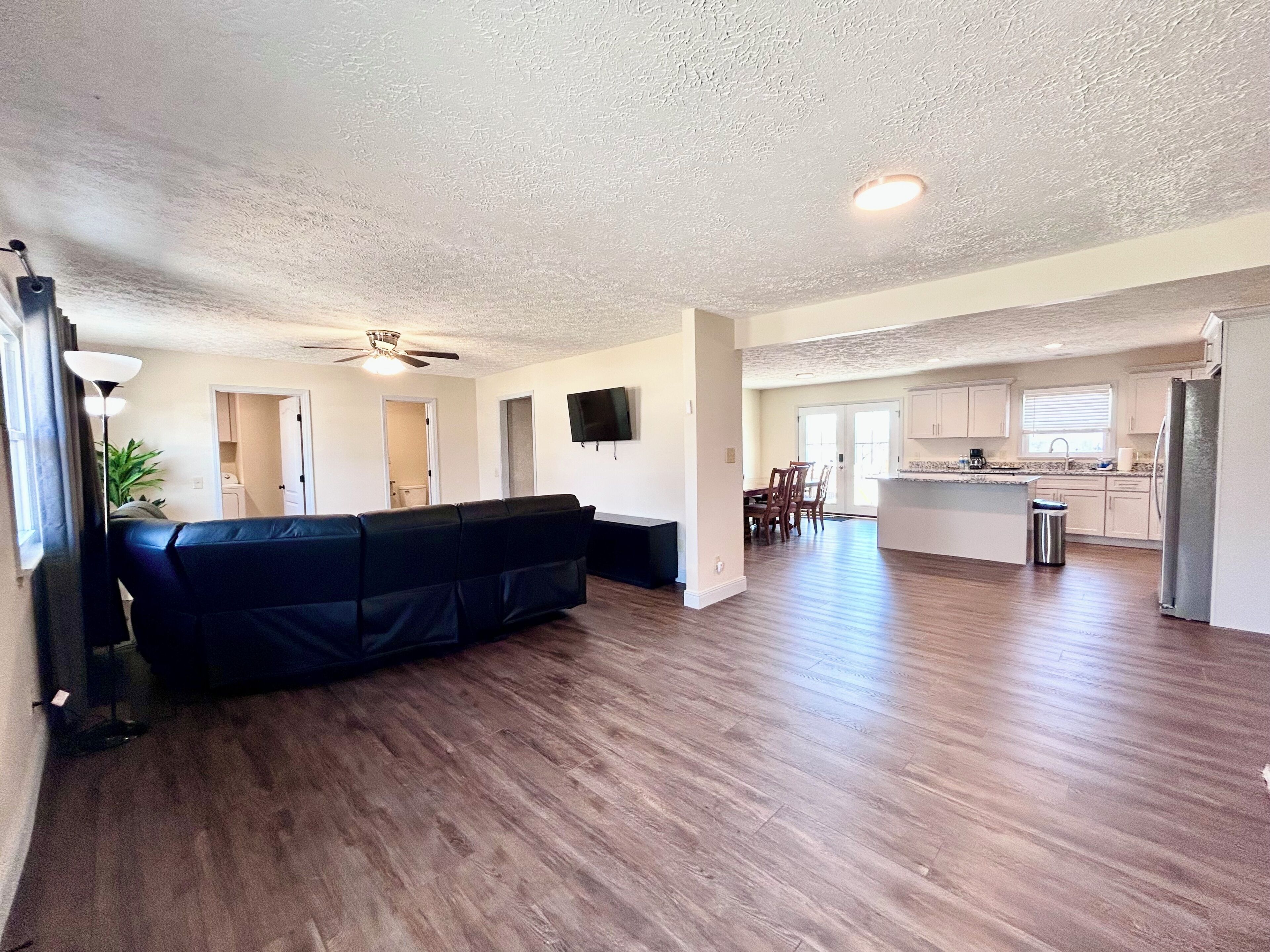 A spacious living area with a dark-colored sofa, wooden flooring, a ceiling fan, and a kitchen area with white cabinets and a stainless steel refrigerator.