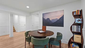 Ein Essbereich mit einem runden Tisch und vier Stühlen, ein Bücherregal mit Büchern und Spielen sowie ein großes Landschaftsbild an der Wand.