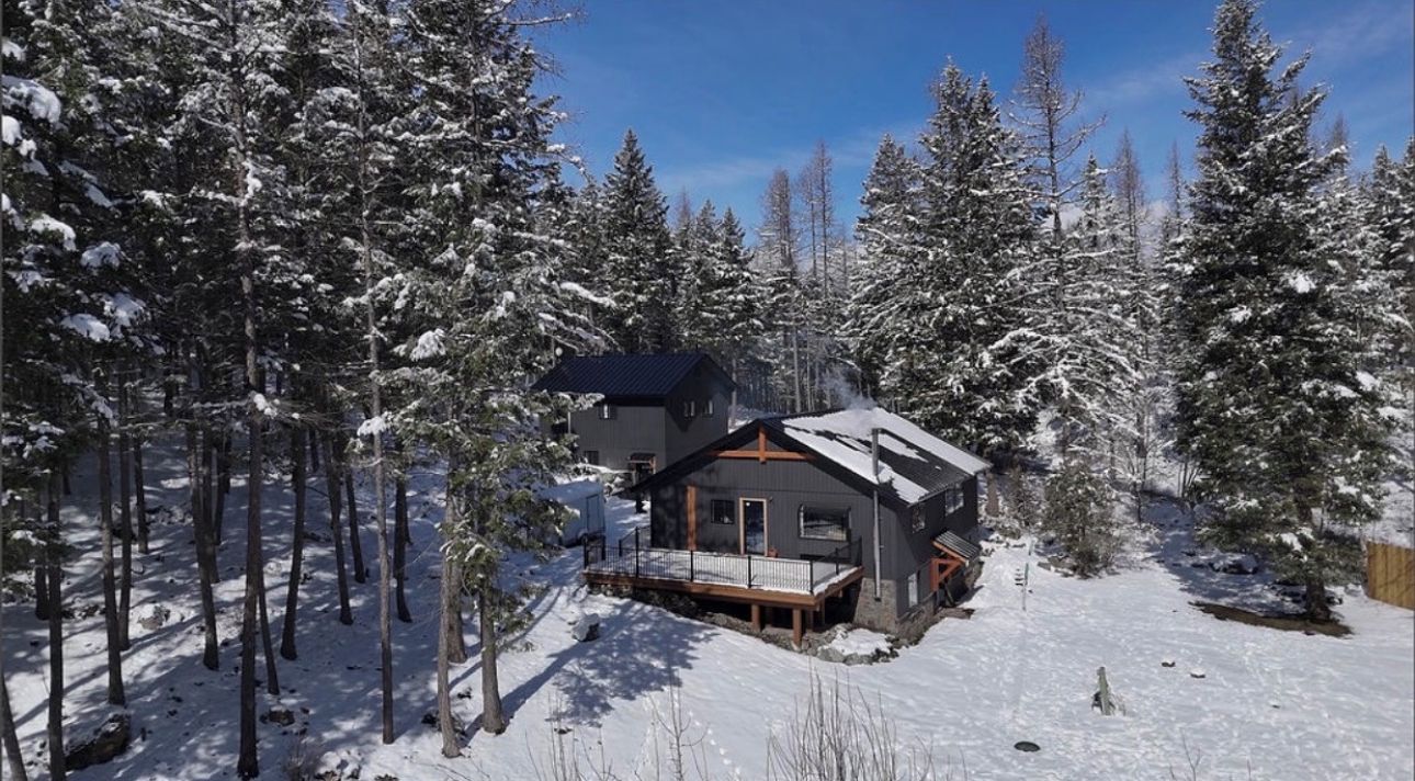 Une cabane entourée d’arbres couverts de neige, avec une terrasse et une grande fenêtre.