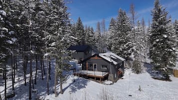 Une cabane entourée d’arbres couverts de neige, avec une terrasse et une grande fenêtre.