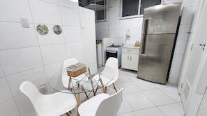 A compact kitchen with white tiles, a stainless steel refrigerator, a gas stove, and a glass dining table with white chairs. - 3 Bedrooms in Copacabana - Near Arpoador (Rio de Janeiro)