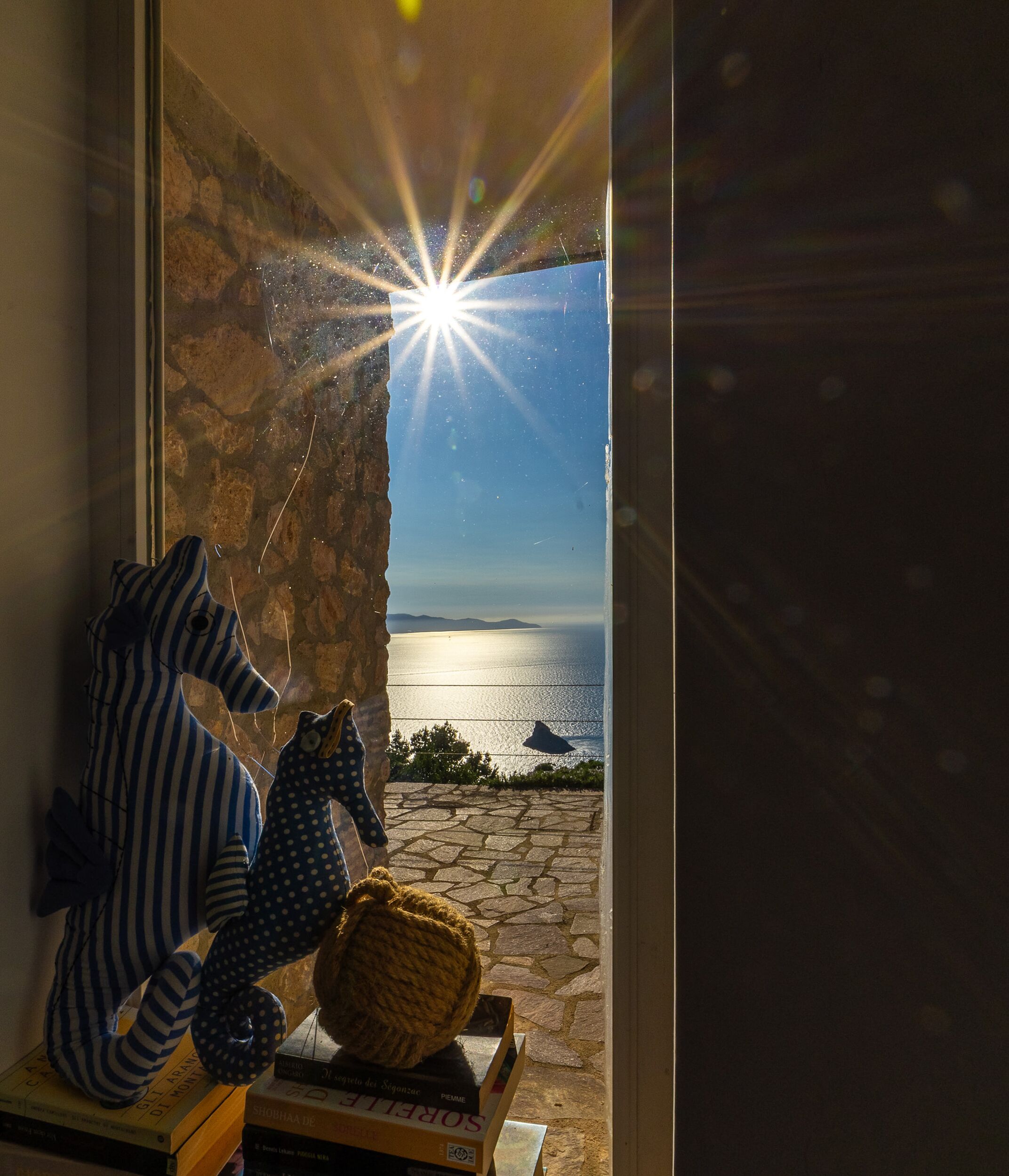 A window with a view of the sea, a stone wall, and stuffed toys on a table.