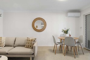 A living room with a sofa, a round table with chairs, a mirror, and an air conditioning unit. - Burleigh Breeze 2‑Bed Beach Walk Retreat (Gold Coast)
