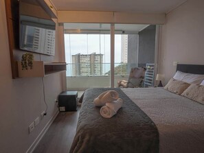 A hotel room with a large bed, a television mounted on the wall, and a view of high-rise buildings through the window. - Costa Home - 7 PAX  & Sunset Arena & Vistas al Mar (Concón)