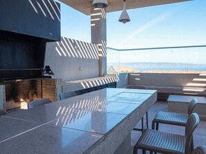 An outdoor patio with a long table and chairs, a grill, and a view of the landscape. - Costa Home - Departamento Vista Valparaíso (Concón)