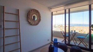 Una habitación con un ventanal grande, un espejo redondo tejido, una escalera y una mesa con dos botellas.