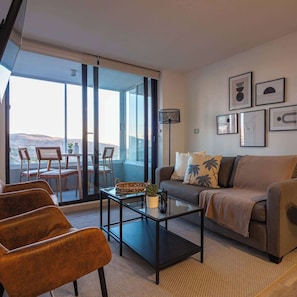 Ein modernes Wohnzimmer mit Sofa, Couchtisch und Sesseln, ein Balkon mit Tisch und Stühlen sowie Kunstwerken an den Wänden.