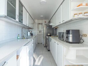 Eine schmale Küche mit weißen Schränken, einer Kaffeemaschine und einem Kühlschrank.