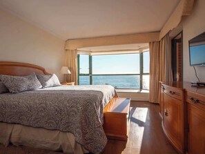 Habitación de hotel con una cama grande, una mesita de noche de madera, un televisor y vista al mar.