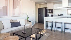 A modern living room with a sofa, coffee table, and bar stools. There is a kitchen area with a refrigerator and stove. - Costa Home 7 PAX - Lujoso - WIFI 1000 MBPS HS (Concón)