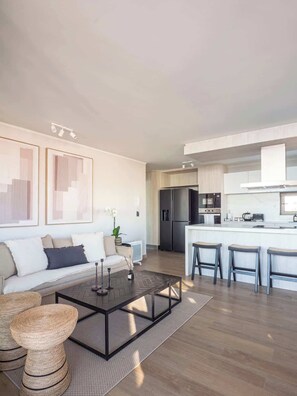 A modern living room with a sofa, coffee table, and bar stools. There is a kitchen area with a refrigerator and stove. - Costa Home 7 PAX - Lujoso - WIFI 1000 MBPS HS (Concón)