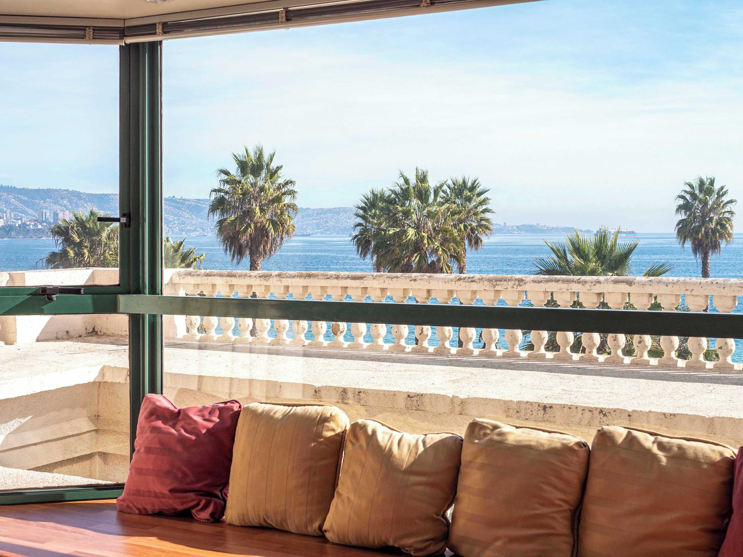 A balcony with a view of the sea and palm trees, a railing, and a sofa with cushions.