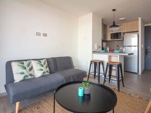 Een moderne woonkamer met een grijze bank, een ronde salontafel en een keuken met een magnetron en een koelkast.