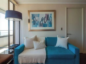 A room with a blue sofa, white cushions, a framed abstract artwork, a floor lamp, and a door. - Costa Home - Departamento Zafiro 1 (Concón)