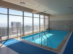 An indoor swimming pool with a view of a cityscape through large windows. - Costa Home - Departamento Zafiro 1 (Concón)
