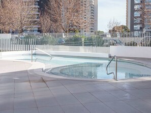Ein Schwimmbecken mit einer gefliesten Sonnenterrasse, umgeben von einem weißen Zaun, mit Gebäuden im Hintergrund.
