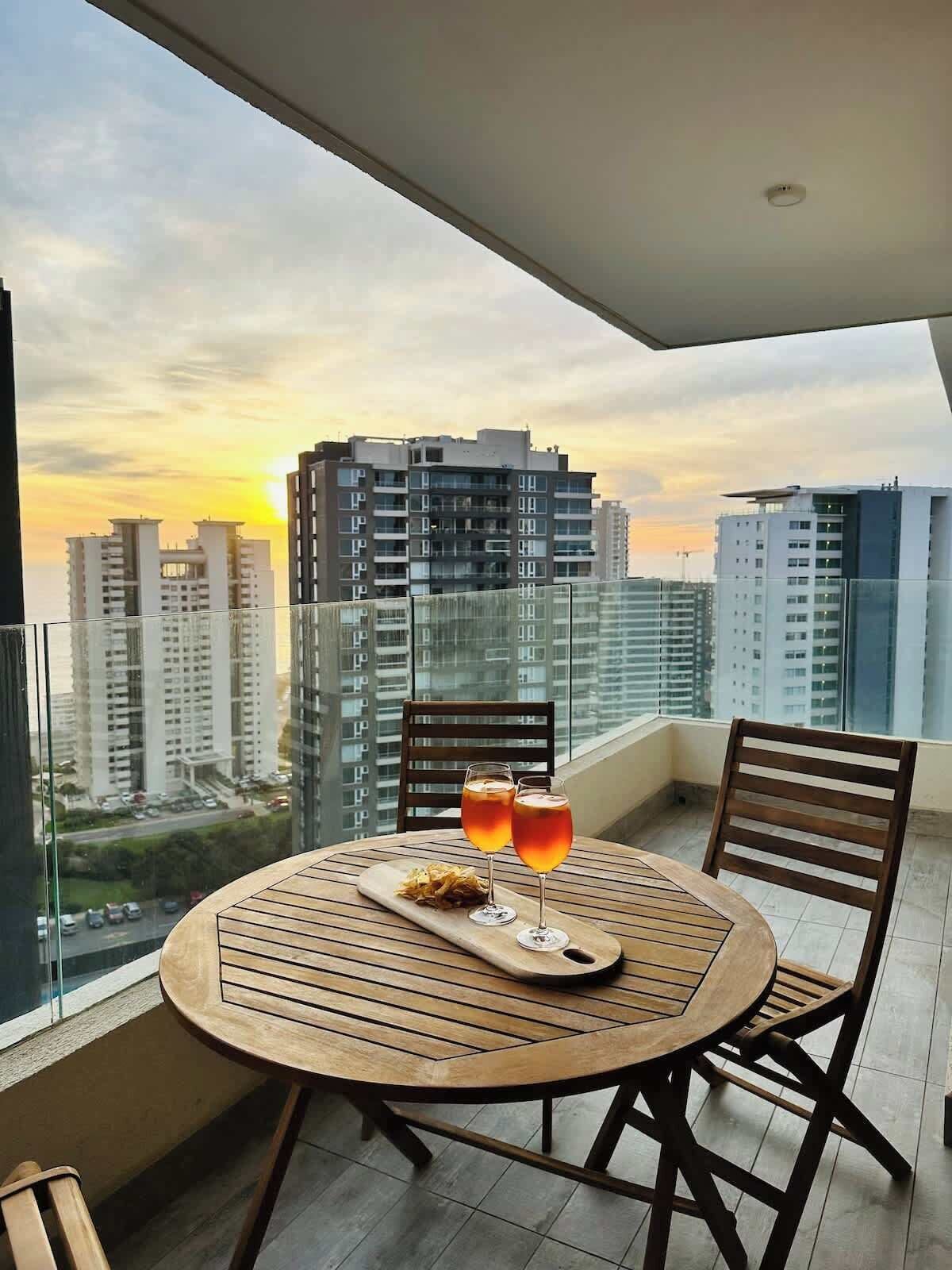 A round wooden table with two glasses of amber liquid, a wooden tray, and two wooden chairs on a balcony with a cityscape view.