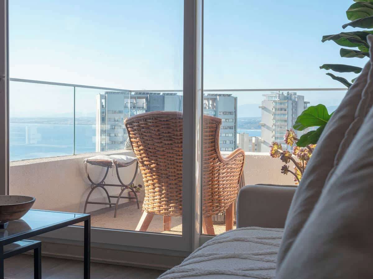Een balkon met rieten stoelen, een tafel en uitzicht op gebouwen en de zee.