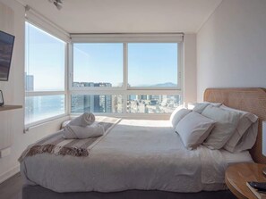 Une chambre avec un grand lit, une table de chevet, une fenêtre donnant sur la ville et un téléviseur fixé au mur.