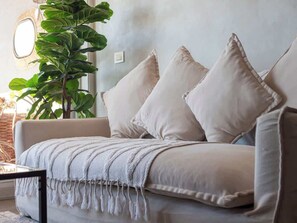 En beige sofa med et vamset pled og tre puder, placeret op ad en væg med en stor grøn plante til venstre.
