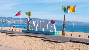 A beachside sign with the word 'VINA' and flags, a palm tree, and a cityscape in the background. - Costa Home - Departamento La Marina (Viña del Mar)