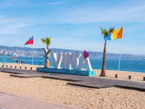 Ein Strandwegweiser mit dem Wort 'VINA' und Flaggen, einer Palme sowie einer Stadtlandschaft im Hintergrund.