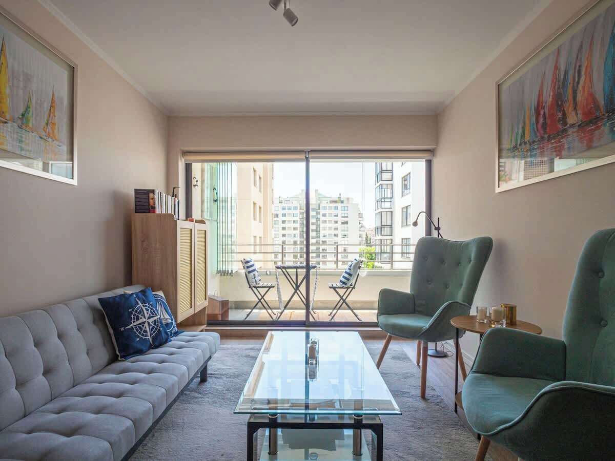 A modern living room with a sofa, armchairs, a glass coffee table, and a balcony with a view of buildings.
