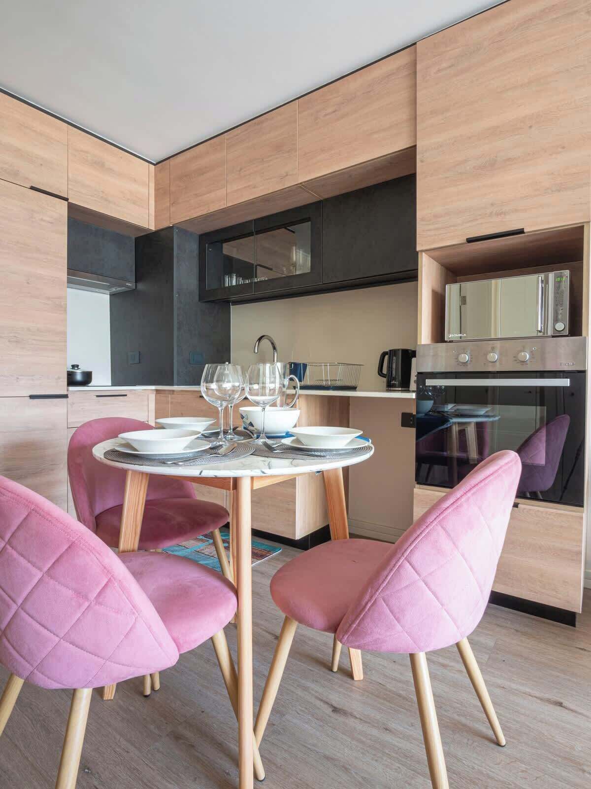 Une cuisine moderne avec des meubles en bois, un four encastré et un coin repas comprenant des chaises à revêtement rose et une table ronde.