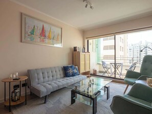 A living room with a sofa, a glass coffee table, a bookshelf, and a view of the city through large windows.