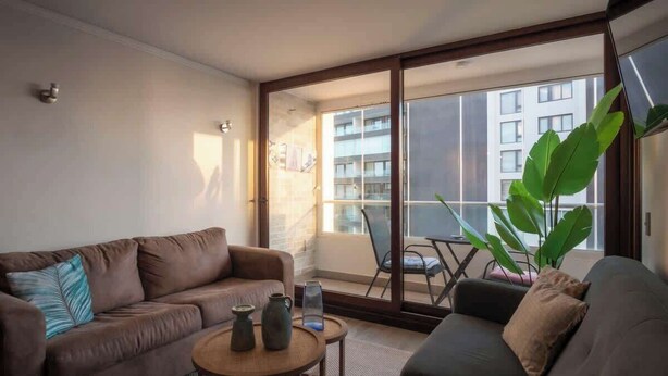 Una sala de estar moderna con un sofá de cuero marrón, una mesa de centro redonda y un ventanal con vistas a un edificio y un balcón.