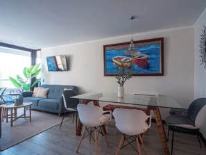 A modern living room with a glass dining table, a sofa, and a painting of boats. - Costa Home - Departamento Eluchans Plaza 1 (Viña del Mar)