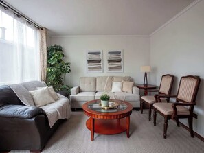 Ein Wohnzimmer mit einer Couch, einem Sessel, einem runden Couchtisch und zwei Stühlen. Eine Pflanze und Kunstwerk sind an der Wand zu sehen.