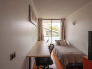 A small, single-room hotel room with a bed, a desk, and a chair. There is a window with curtains, a wall-mounted light, and a door.