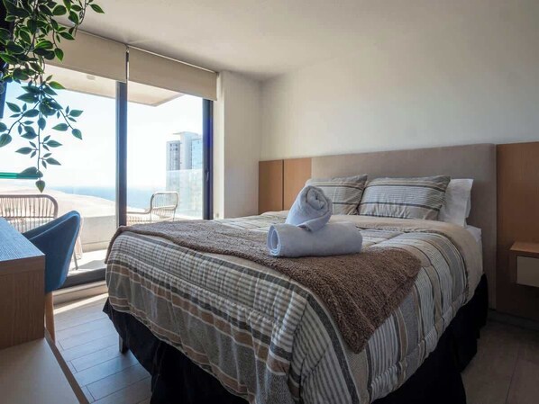 Habitació d'hotel amb un llit, una cadira, una taula petita i vistes a la ciutat a través de grans finestres.