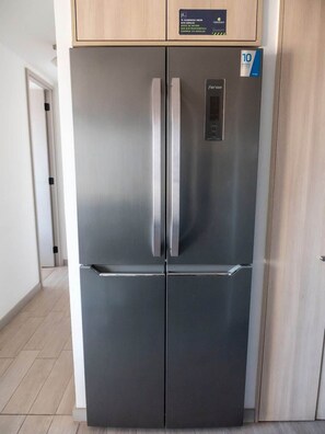 A stainless steel refrigerator with a digital display, a wooden cabinet above, and a tiled floor. - Costa Home - Dpto EcoCostas Concón (Concón)