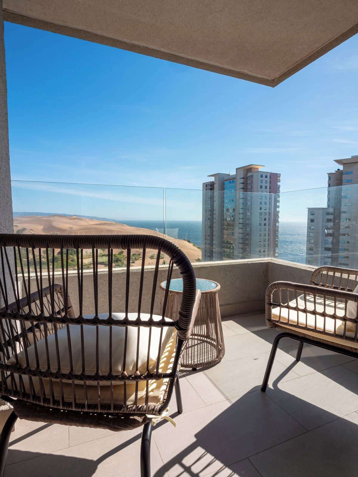 A balcony with two wicker chairs and a small round table, overlooking a cityscape and a desert.