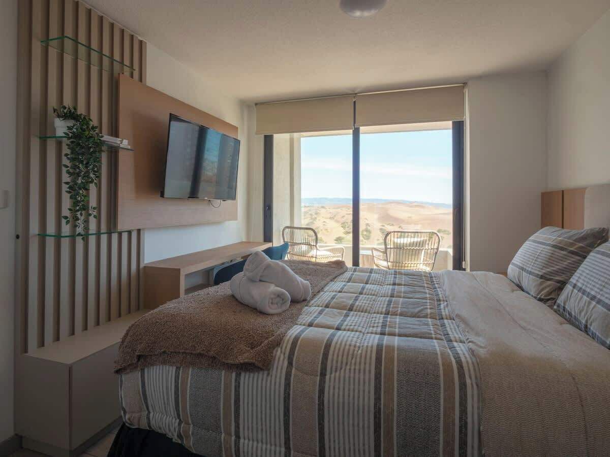 A bedroom with a bed, a TV, a plant, a chair, and a view of the desert.