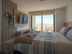 A bedroom with a bed, a TV, a plant, a chair, and a view of the desert. - Costa Home - Dpto EcoCostas Concón (Concón)