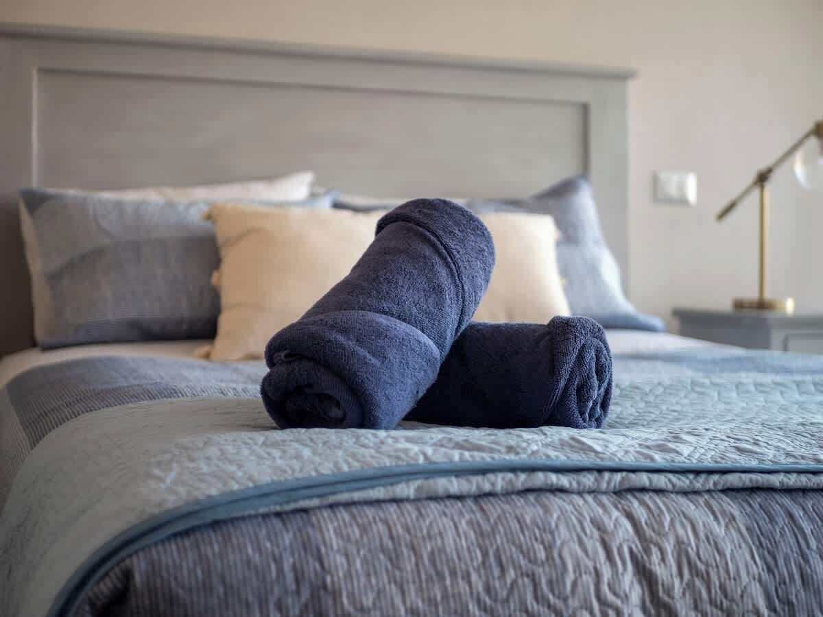 Een net opgemaakt bed met een grijs hoofdeinde, een blauwe quilt en twee opgerolde handdoeken.