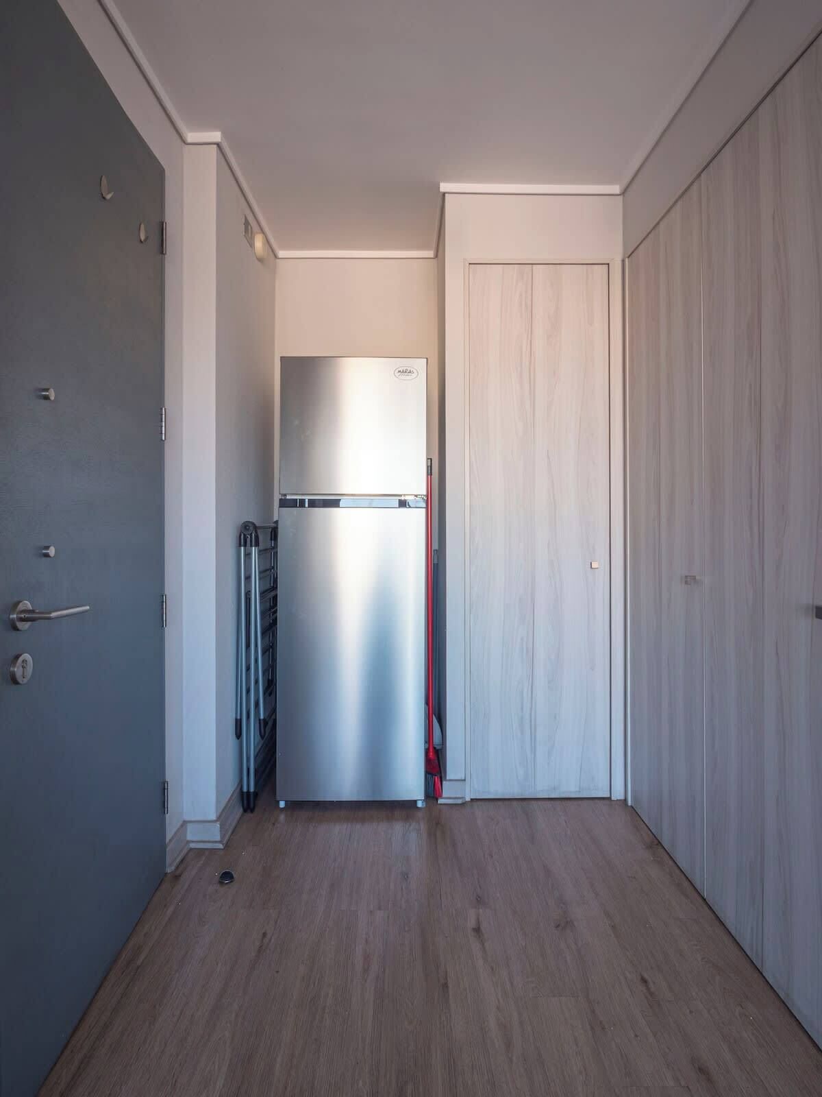 A narrow hallway with a stainless steel refrigerator, a door with a handle, and a wooden floor.