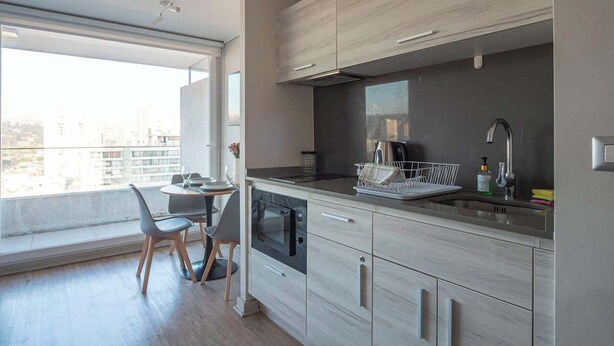 A modern kitchen with a built-in oven, a dining area with a table and chairs, and a large window offering a city view. - Apartamento 1 Habitación Quinta Vergara (Viña del Mar)
