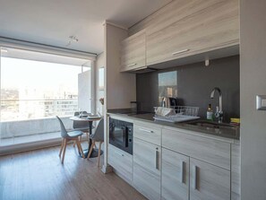 A modern kitchen with a built-in oven, a dining area with a table and chairs, and a large window offering a city view. - Apartamento 1 Habitación Quinta Vergara (Viña del Mar)