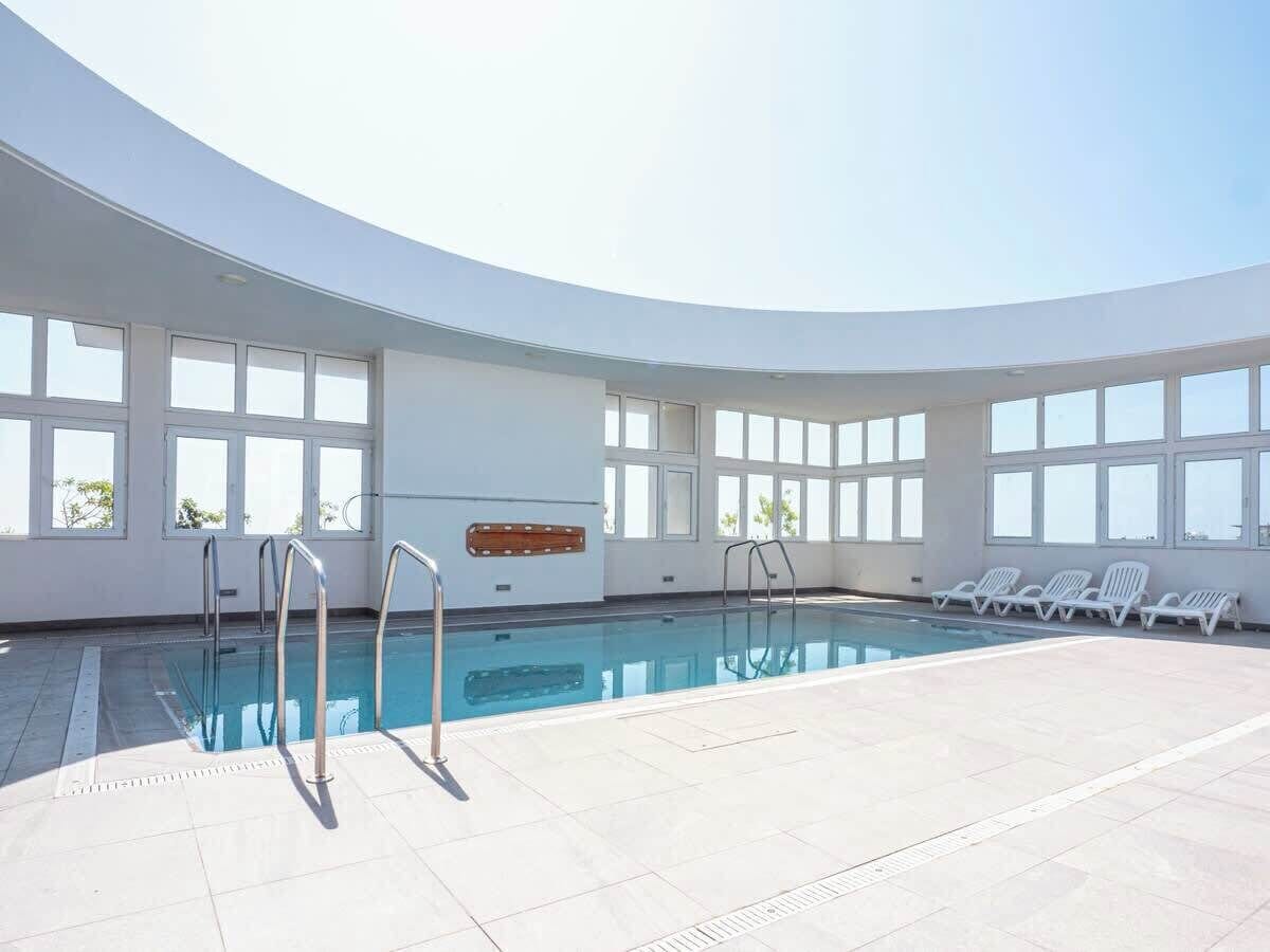 Une grande piscine intérieure aux parois en verre transparent, dotée d’une verrière et bordée d’une rangée de chaises longues.