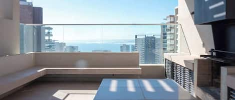 A modern balcony with a view of the sea, a seating area, and a built-in grill.