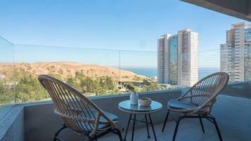 A balcony with two wicker chairs and a small table, overlooking a cityscape and hills.