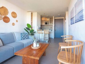 A modern living room with a sofa, wooden coffee table, and wicker chairs. A kitchen is visible in the background with bar stools.