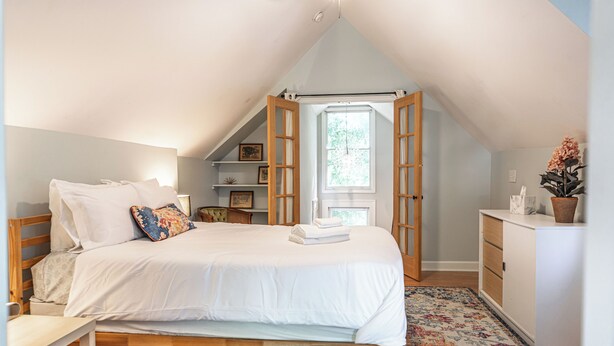 Une chambre à coucher avec un lit, une table de chevet, une tête de lit en bois, une fenêtre ouverte, un ventilateur de plafond et une plante en pot.