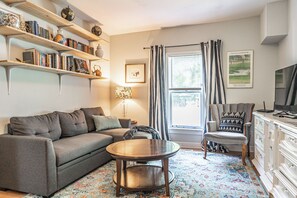 A living room with a sofa, armchair, coffee table, bookshelf, lamp, and a TV. - NEW! Pastry Townhouse - Steps to historic Southern Pines! (Southern Pines)